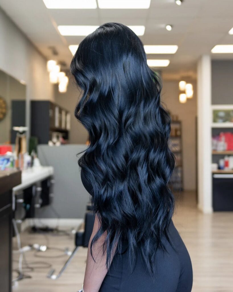 Long wavy black-blue hair shown from behind inside a bright salon
