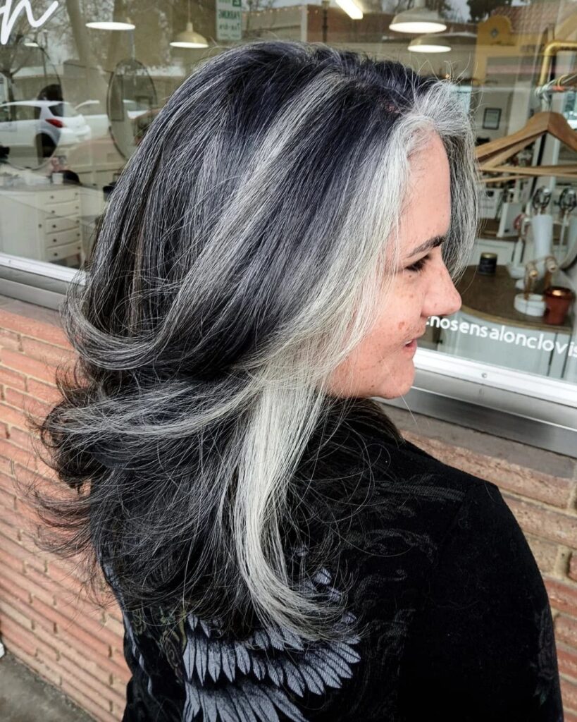 Layered dark hair with a bold silver face-framing streak beside a salon window