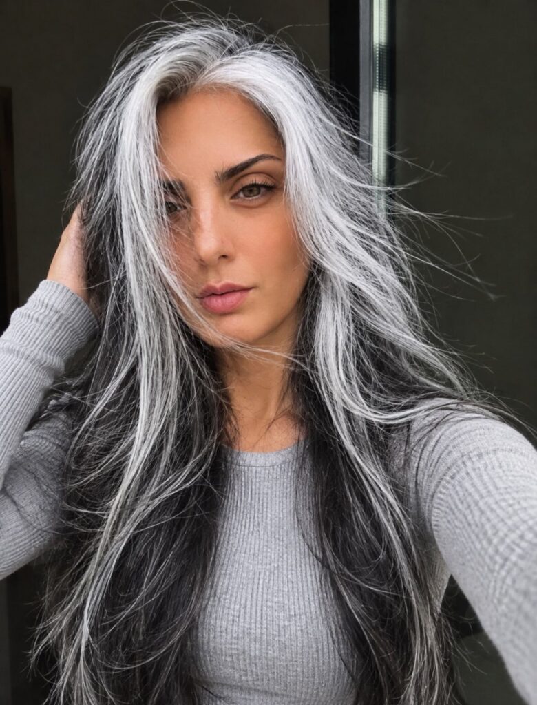 Woman with long wavy salt and pepper hair wearing a light gray ribbed top