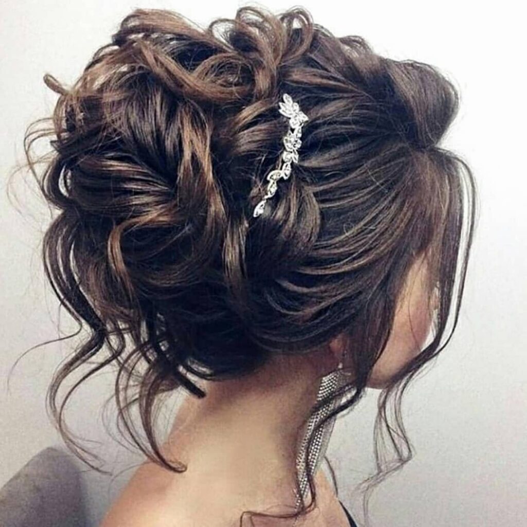 Brown curly bridal updo with loose tendrils and a sparkling floral hairpiece